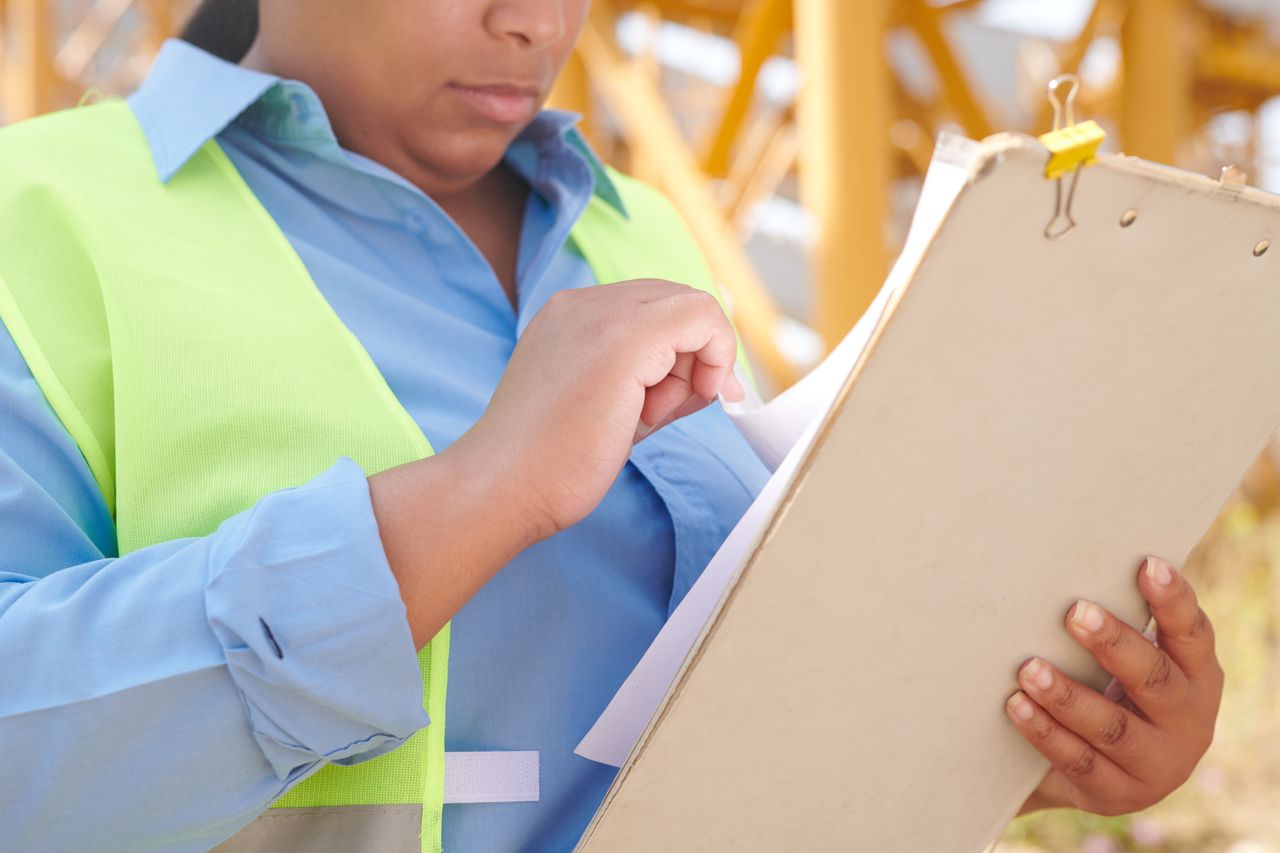 A female engineer in safety gear reviewing documents on a clipboard at a construction site.